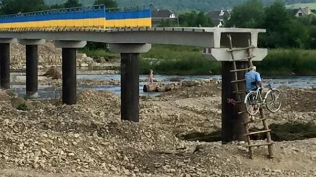 Люди по стремянке с велосипедами на спине вылезают на недостроенный мост Люди по стремянке с велосипедами на спине вылезают на недостроенный мост