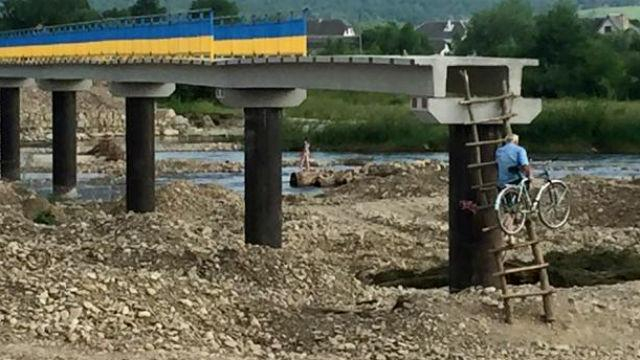 Люди по стремянке с велосипедами на спине вылезают на недостроенный мост